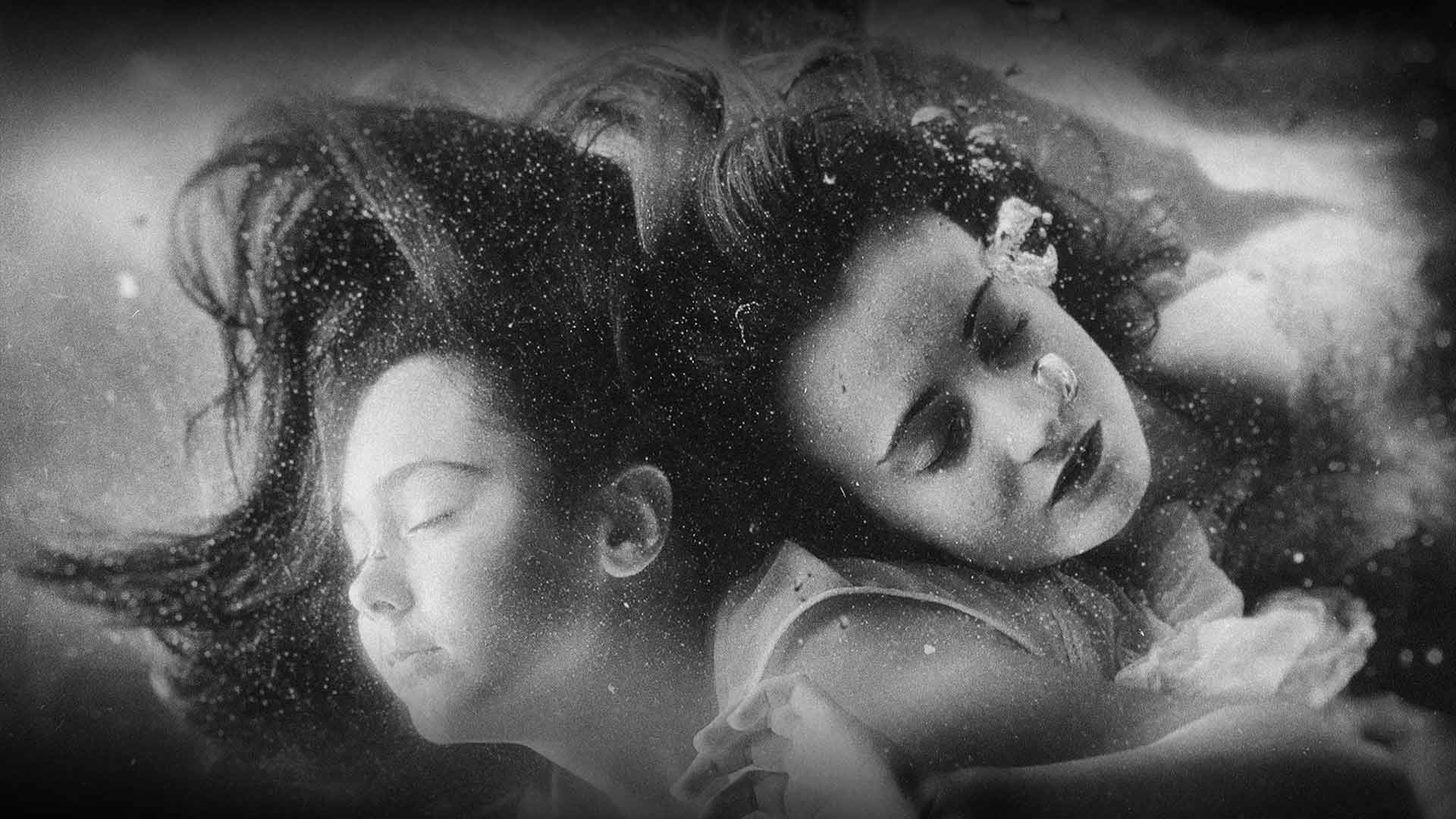 sepia-toned underwater photograph featuring two young women with their eyes closed, floating in a dreamlike, suspended state.